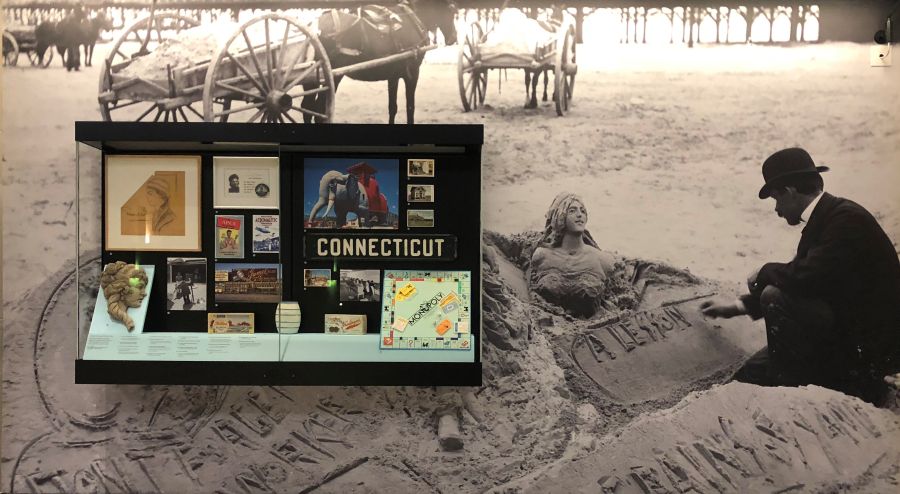 Photograph of Innovation and Invention exhibit case with photographs and artifacts depicting the sand artists, a sand sculpture with concrete, Sara Spencer Washington's business, Monopoly board game with pieces, Lucy the Elephant, saltwater taffy boxes and a coin bank.